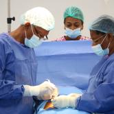 Two Haitian surgeons in full PPE, one male and one female, begin operating on a patient. Another clinician observes from behind a blue drape.