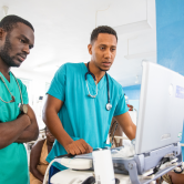 Two Haitian men doctors wearing light blue scrub tops look at a sonogram machine in an emergency room.
